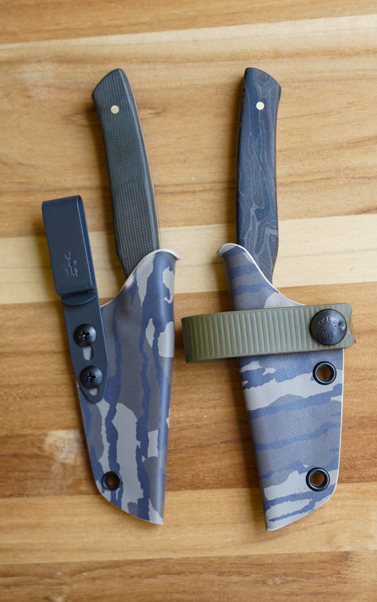 Two of the best fixed-blade knives with blue-gray camo sheaths and dark wood handles, displayed on a light brown wooden surface.