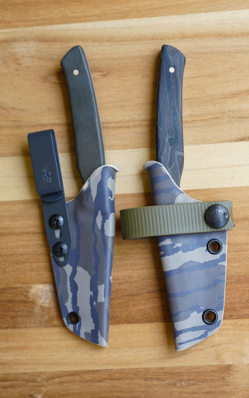 Two of the best fixed-blade knives with blue-gray camo sheaths and dark wood handles, displayed on a light brown wooden surface.